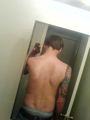 man facing away from camera.  The photo is in a mirror.  One can see both his scoliosis scars from surgery and his elaborate arm tattoo.