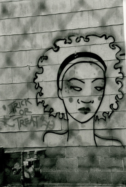 graffiti of a woman's head, face, with "trick or treat" written next to it...photo taken from behind a wire fence, so the image looks fenced-in