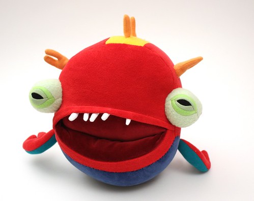 A round, red monster with a blue underside. It has two extending eyes, also circular, with light green irises, and several little orange horns on its head. Its maroon mouth is big and has only five little white teeth, all in the top row. The crease in the back of the mouth that leads to the pocket is visible.