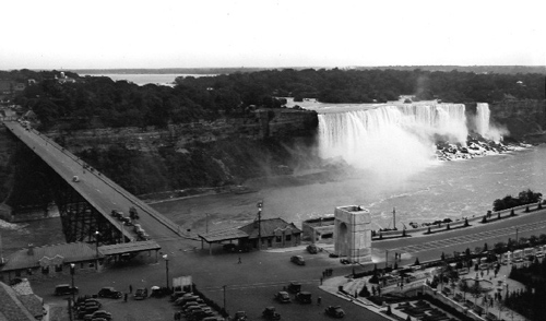 Memorial Arch (centre right) with Honeymoon Bridge and America Falls, 1937. This view of the Memorial Arch shows the Falls View or Honeymoon Bridge bringing American tourists directly to its feet.  In January 1938, the bridge collapsed, and was replaced with the new Rainbow Bridge, considerably upstream.  