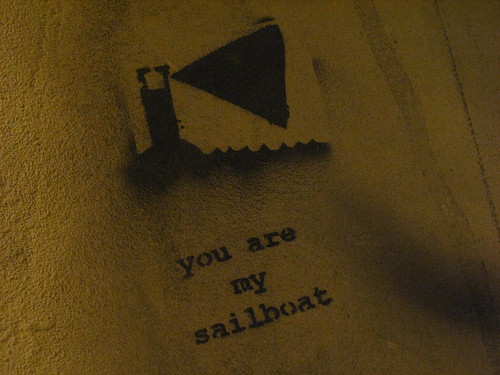stencil graffiti of a lighthouse shining a triangle of light out over water, with the words, 'you are my sailboat'