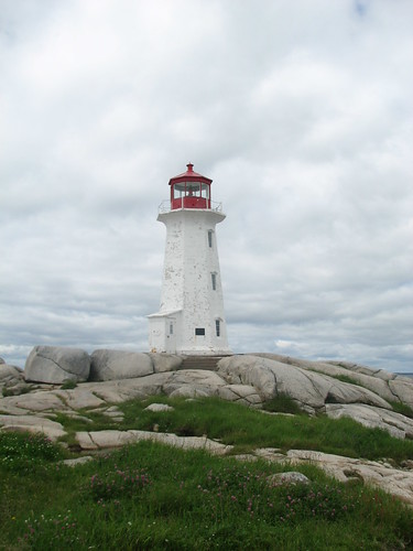 The famous Peggy's Cove lighthouse. It's quite tall, and white, with the actual light casing in red. There are three windows going up on the side. It really needs a new coat of paint. In the foreground is grass and rocks leading up the lighthouse. In the background is just cloud. There's a storm coming in. Posted by Anna Overseas, on Flickr, description written by Anna