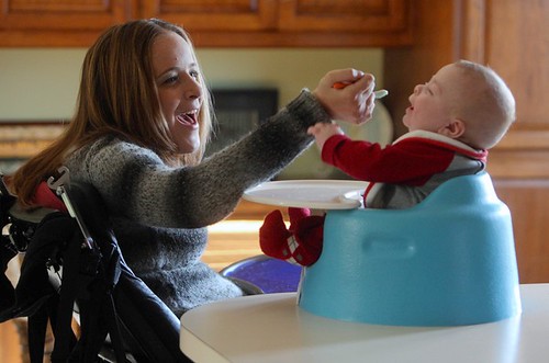 A smiling Kaney O'Neill, sitting in a wheelchair, spoon feeds her baby who is sitting in a Bumbo type seat. 