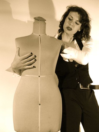 A person with medium-length curly hair, wearing black pants with a belt, a white shirt, a black waistcoat, lipstick and nail polish. They are holding onto a headless, unclothed mannequin as though working with it.