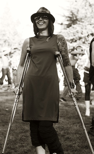 A tall slender woman wearing a slip dress and an awesome hat.  She's got tattoos on one arm, and is using crutches.  She's grinning.