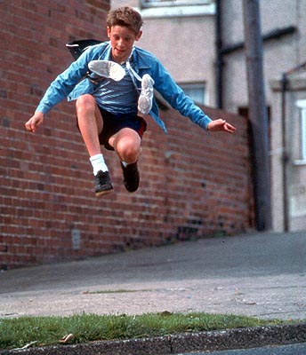 Billy Elliot, a white boy wearing a blue jacket, jumps in the air. Ballet shoes hang around his neck.
