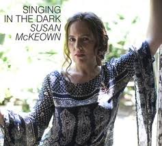 Susan McKeown, a white woman, wears a printed dress and looks at the camera. Her album cover reads Singing in the Dark in dark text