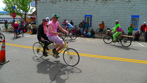 Right side view of 2 cyclists, riding on a double bike, down a city street with a building in the background