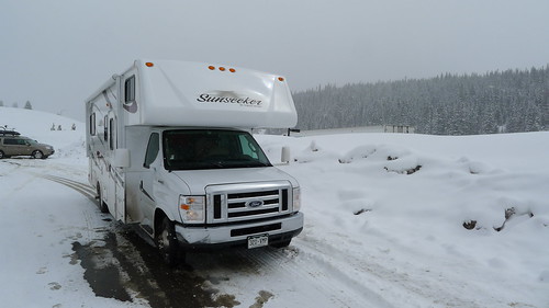 Front, right side view of a motorhome, driving on a snow covered road, with snowy trees in the background
