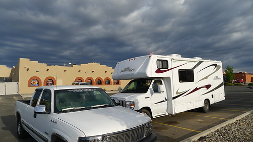 A white pickup truck next to a motorhome, on a paved parking lot with a building in the background and clouds in the sky