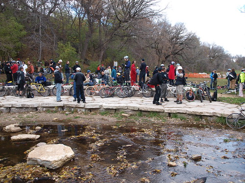 Side view of a stone walking path with people and bikes on it, in front small pool of water, with trees in background