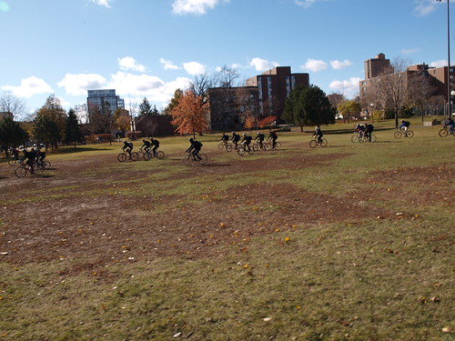 Left side view of cyclists riding bikes across a grass field with buildings behind it and blue sky above