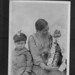 Seated woman holding an infant in a cradle board, with a young boy sitting next to them... / Femme assise tenant un bébé dans un porte-bébé; un jeune garçon est assis à côté d’eux...